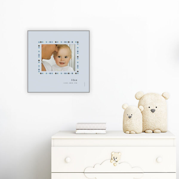viser kvadratisk fødselstavle med et foto omkranset af en soed kant af blomster i blaa farver. Dertil fornavn og foedselsdata. Graa baggrund-Designet af www.plakatcompagniet.dk. plakat haenger paa boernevaerelse over hvid kommode