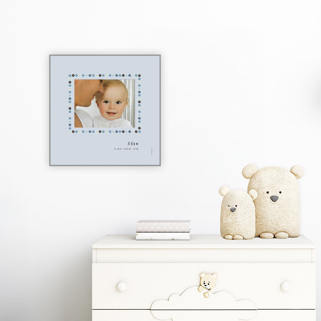 viser kvadratisk fødselstavle med et foto omkranset af en soed kant af blomster i blaa farver. Dertil fornavn og foedselsdata. Graa baggrund-Designet af www.plakatcompagniet.dk. plakat haenger paa boernevaerelse over hvid kommode