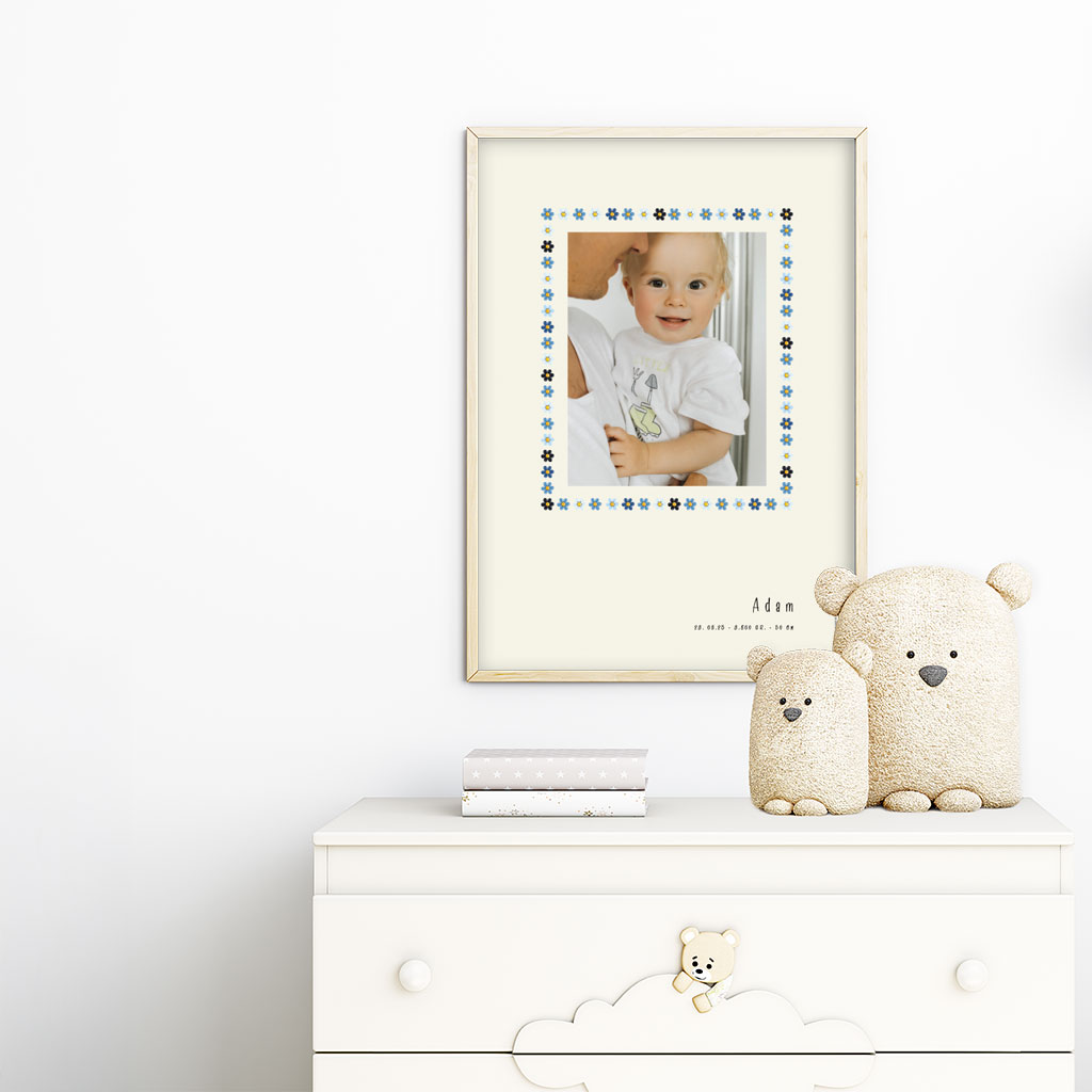 viser rektangulaer fødselstavle med et foto omkranset af en soed kant af blomster i blaa farver. Dertil fornavn og foedselsdata. Beige baggrund. Designet af www.plakatcompagniet.dk. plakat haenger paa boernevaerelse over hvid kommode
