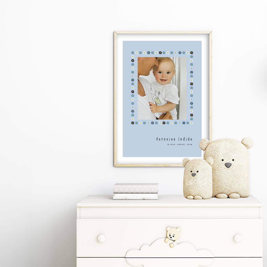 viser rektangulaer fødselstavle med et foto omkranset af en soed kant af blomster i blaa farver. Dertil fornavn og foedselsdata. Blaa baggrund og hvid kant. Designet af www.plakatcompagniet.dk. plakat haenger paa boernevaerelse over hvid kommode
