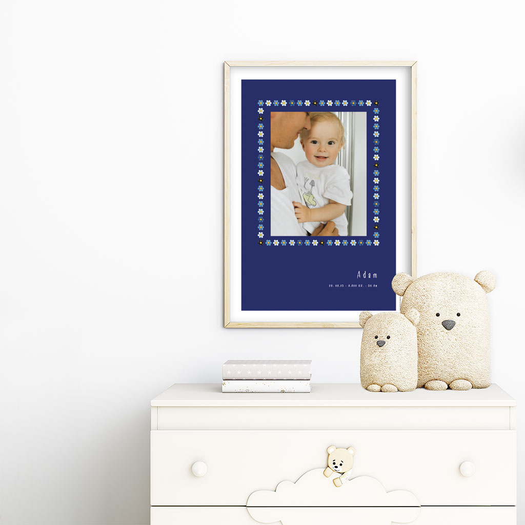 viser rektangulaer fødselstavle med et foto omkranset af en soed kant af blomster i blaa farver. Dertil fornavn og foedselsdata. Moerkeblaa baggrund og hvid kant. Designet af www.plakatcompagniet.dk. plakat haenger paa boernevaerelse over hvid kommode