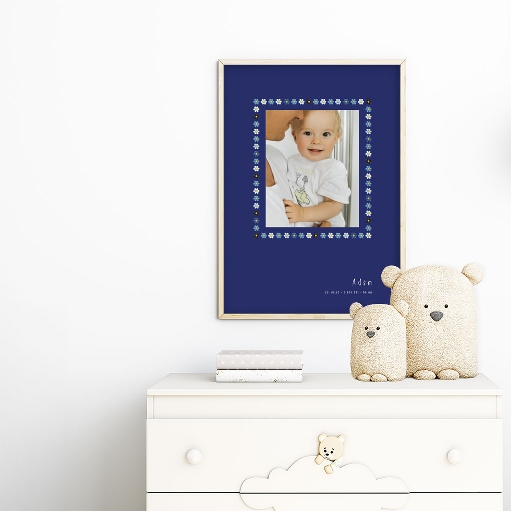 viser rektangulaer fødselstavle med et foto omkranset af en soed kant af blomster i blaa farver. Dertil fornavn og foedselsdata. Moerkeblaa baggrund. Designet af www.plakatcompagniet.dk. plakat haenger paa boernevaerelse over hvid kommode