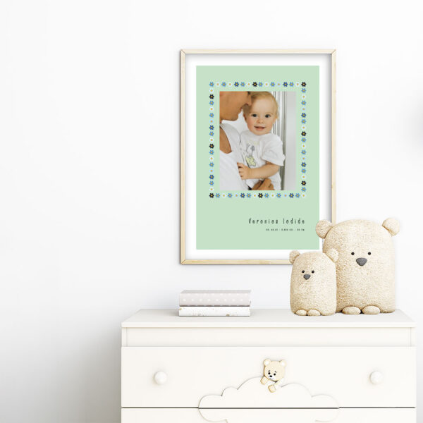 viser rektangulaer fødselstavle med et foto omkranset af en soed kant af blomster i blaa farver. Dertil fornavn og foedselsdata. Mint baggrund og hvid kant. Designet af www.plakatcompagniet.dk. plakat haenger paa boernevaerelse over hvid kommode