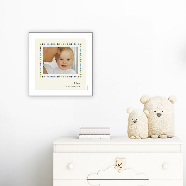 viser kvadratisk fødselstavle med et foto omkranset af en soed kant af blomster i blaa farver. Dertil fornavn og foedselsdata. Beige baggrund og hvid kant. Designet af www.plakatcompagniet.dk. plakat haenger paa boernevaerelse over hvid kommode