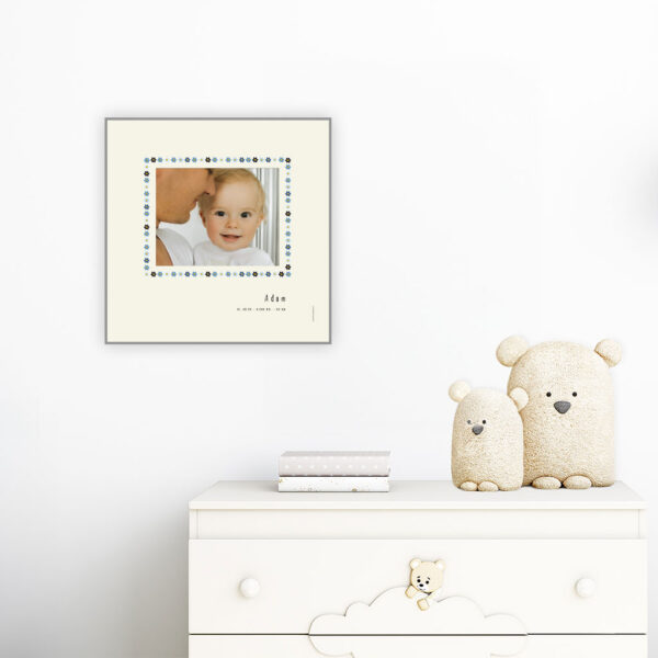 viser kvadratisk fødselstavle med et foto omkranset af en soed kant af blomster i blaa farver. Dertil fornavn og foedselsdata. Beige baggrund-Designet af www.plakatcompagniet.dk. plakat haenger paa boernevaerelse over hvid kommode