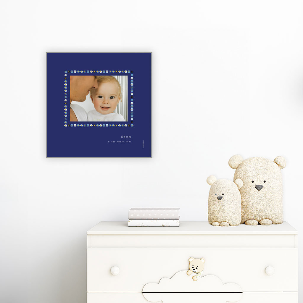 viser kvadratisk fødselstavle med et foto omkranset af en soed kant af blomster i blaa farver. Dertil fornavn og foedselsdata. Moerkeblaa baggrund-Designet af www.plakatcompagniet.dk. plakat haenger paa boernevaerelse over hvid kommode