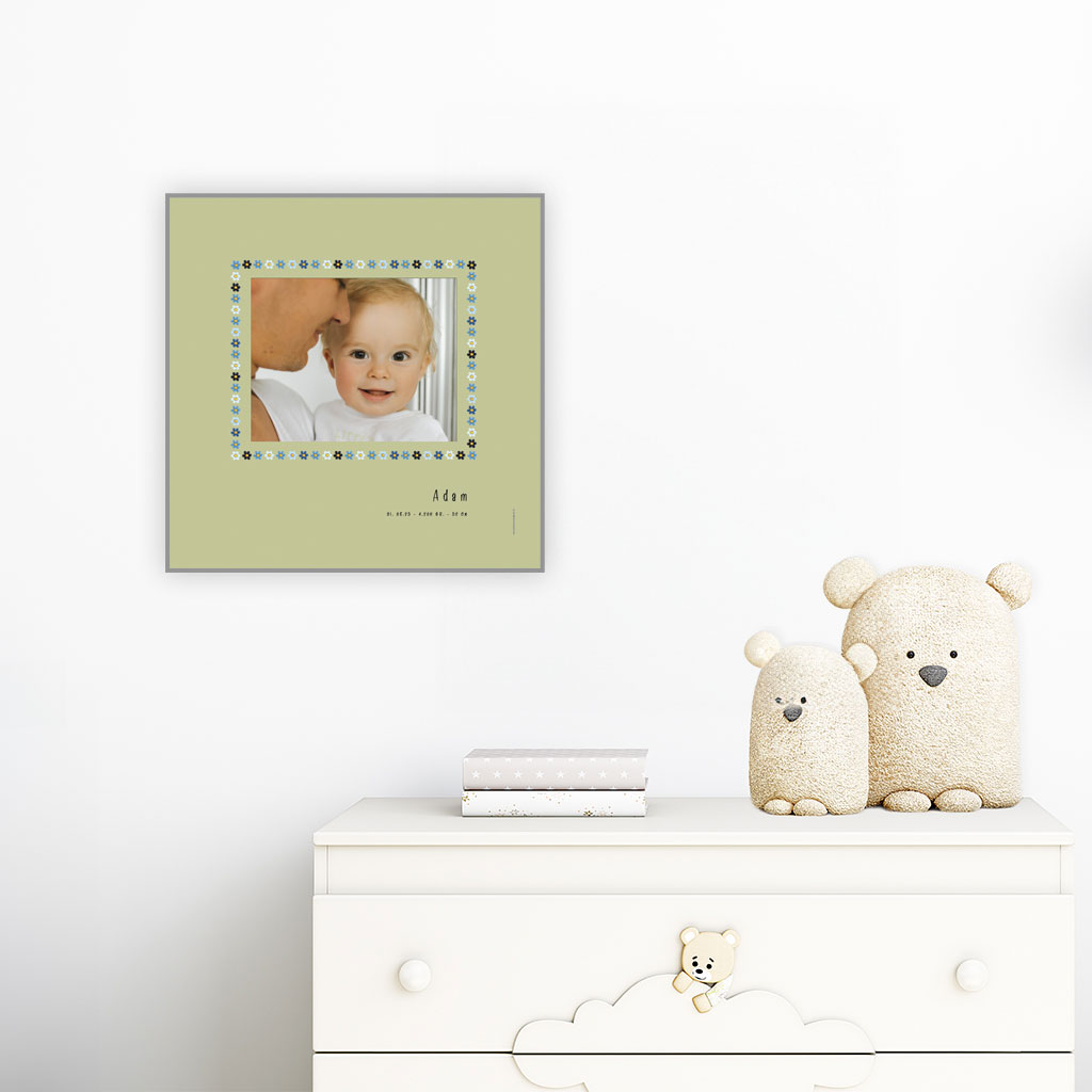 viser kvadratisk fødselstavle med et foto omkranset af en soed kant af blomster i blaa farver. Dertil fornavn og foedselsdata. Groen baggrund-Designet af www.plakatcompagniet.dk. plakat haenger paa boernevaerelse over hvid kommode