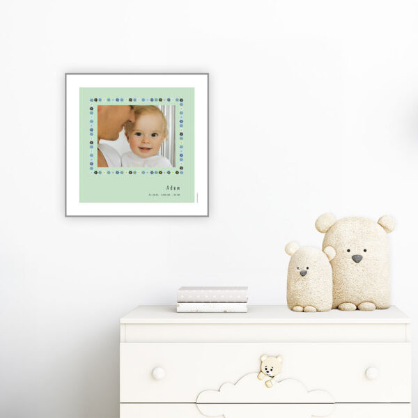 viser kvadratisk fødselstavle med et foto omkranset af en soed kant af blomster i blaa farver. Dertil fornavn og foedselsdata. Mint baggrund og hvid kant. Designet af www.plakatcompagniet.dk. plakat haenger paa boernevaerelse over hvid kommode