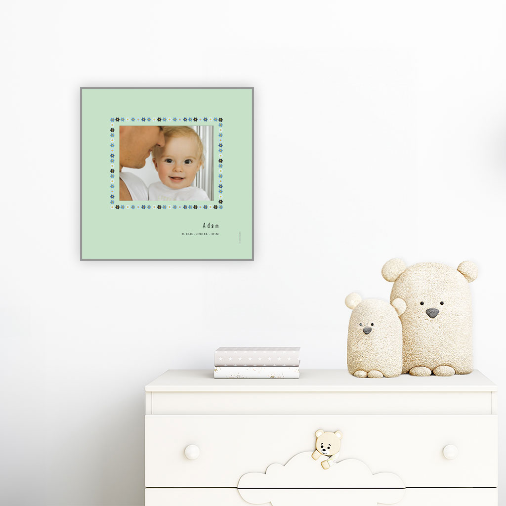 viser kvadratisk fødselstavle med et foto omkranset af en soed kant af blomster i blaa farver. Dertil fornavn og foedselsdata. Mint baggrund-Designet af www.plakatcompagniet.dk. plakat haenger paa boernevaerelse over hvid kommode