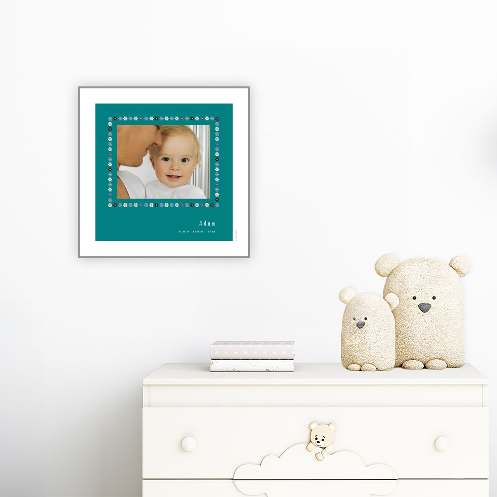 viser kvadratisk fødselstavle med et foto omkranset af en soed kant af blomster i blaa farver. Dertil fornavn og foedselsdata. Teal baggrund og hvid kant. Designet af www.plakatcompagniet.dk. plakat haenger paa boernevaerelse over hvid kommode
