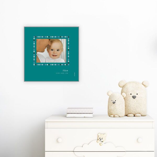 viser kvadratisk fødselstavle med et foto omkranset af en soed kant af blomster i blaa farver. Dertil fornavn og foedselsdata. Teal baggrund-Designet af www.plakatcompagniet.dk. plakat haenger paa boernevaerelse over hvid kommode
